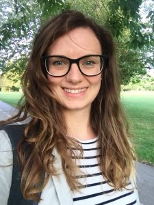 A photograph of Joanna Hastwell smiling for the camera. Joanna is outside in a green space and has long brown hair, wearing a black and white striped shirt and black glasses