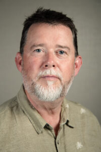A headshot of Dr John Harding, a white man with short dark hair and a grey beard wearing a beige shirt.